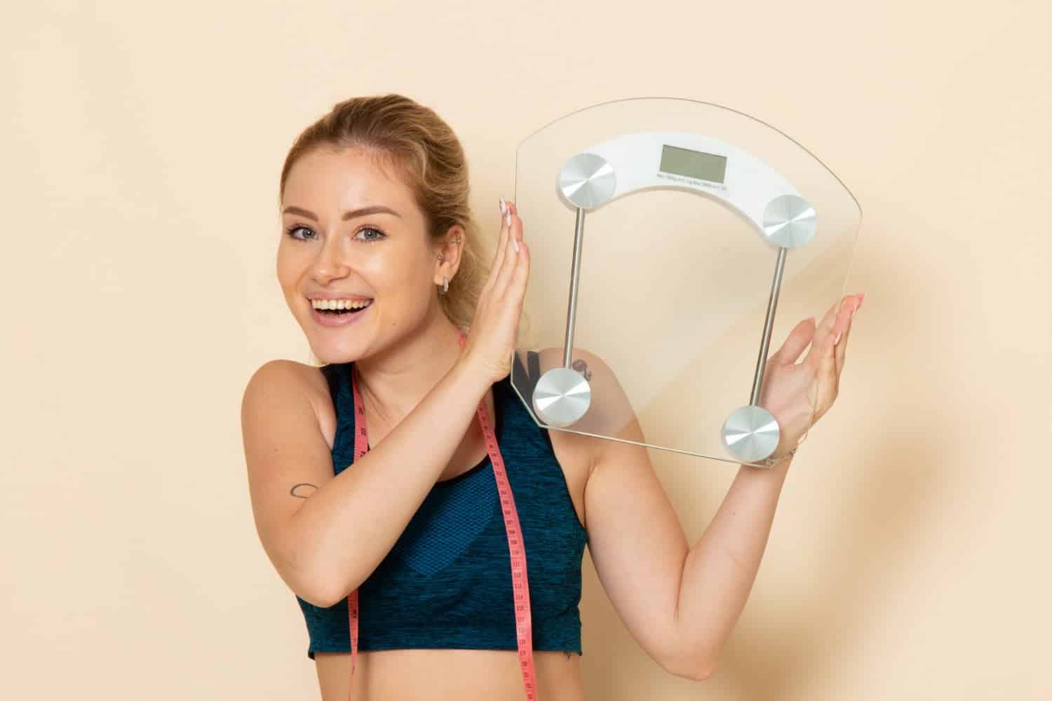 10 benefícios ao Perder Aquele Peso Extra 1 front view young female in sport outfit holding scales on white wall fit body sport beauty health