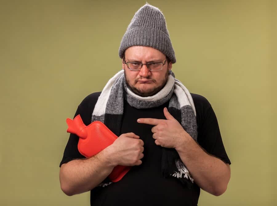 triste homem doente de meia idade usando chapeu de inverno e lenco segurando e apontando para uma bolsa de agua quente isolada na parede verde oliva 141793 127642